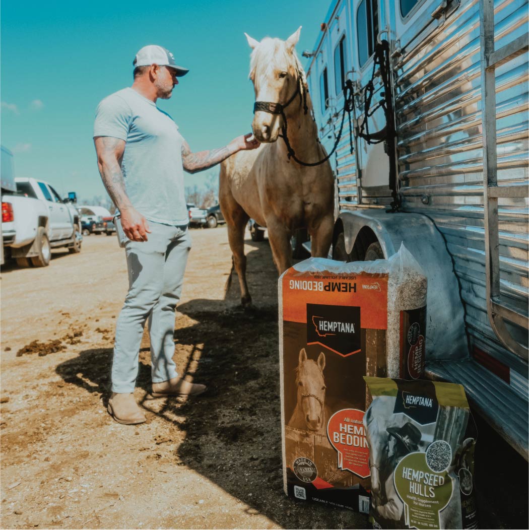 A man standing with a horse tied to a trailer, a 70 L bag of Hemptana Hemp Bedding, and an 8 lb. bag of Hemptana Hempseed Hulls.